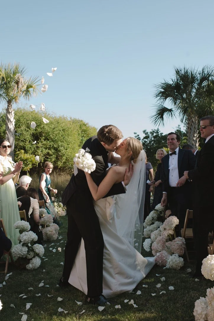 A couple kissing at their outdoor wedding ceremony, surrounded by friends and family, with white floral decorations and palm trees.