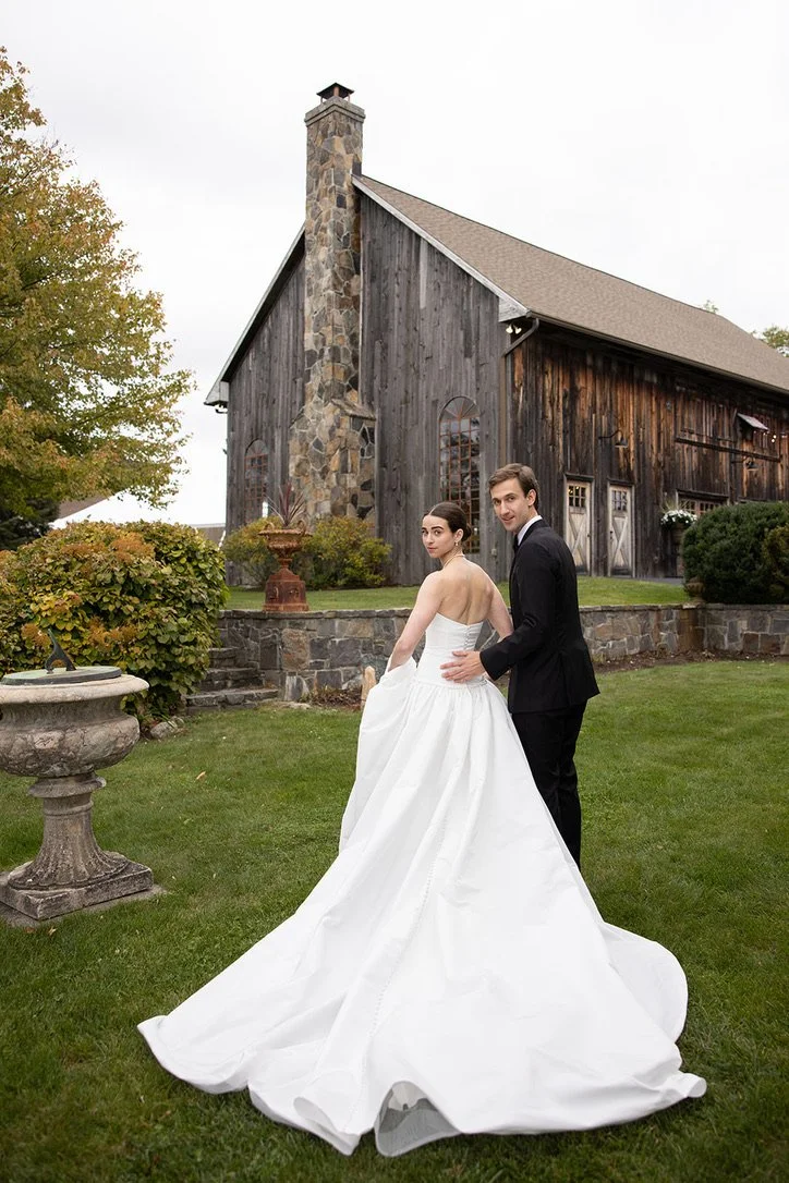 A bride and groom standing on a well-manicured lawn in front of a rustic barn, with the bride wearing a white wedding gown and the groom in a black tuxedo.