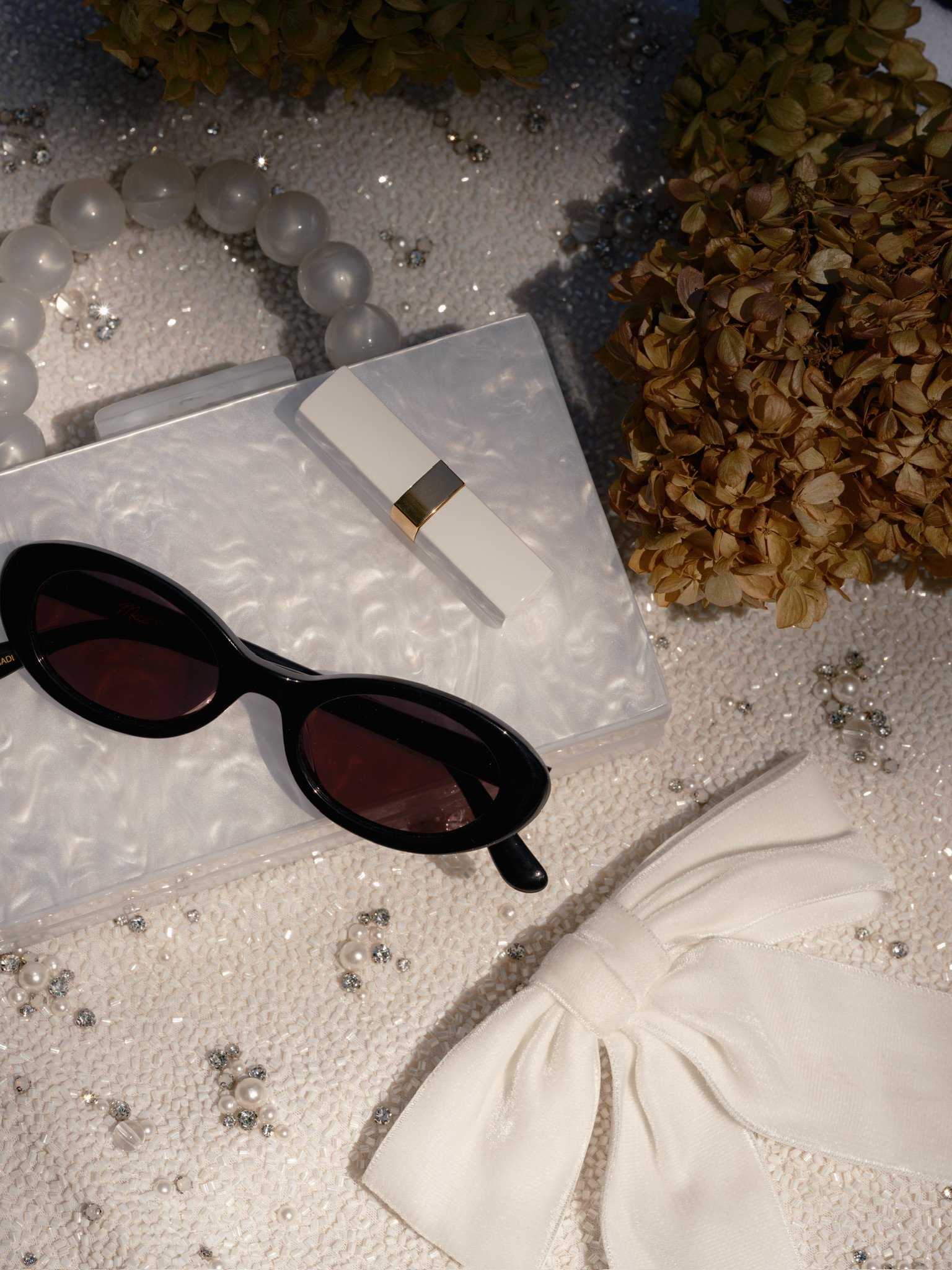 Accessories including black sunglasses, a white lipstick, a white bow, pearl jewelry, and dried flowers on a textured surface decorated with small beads and rhinestones.