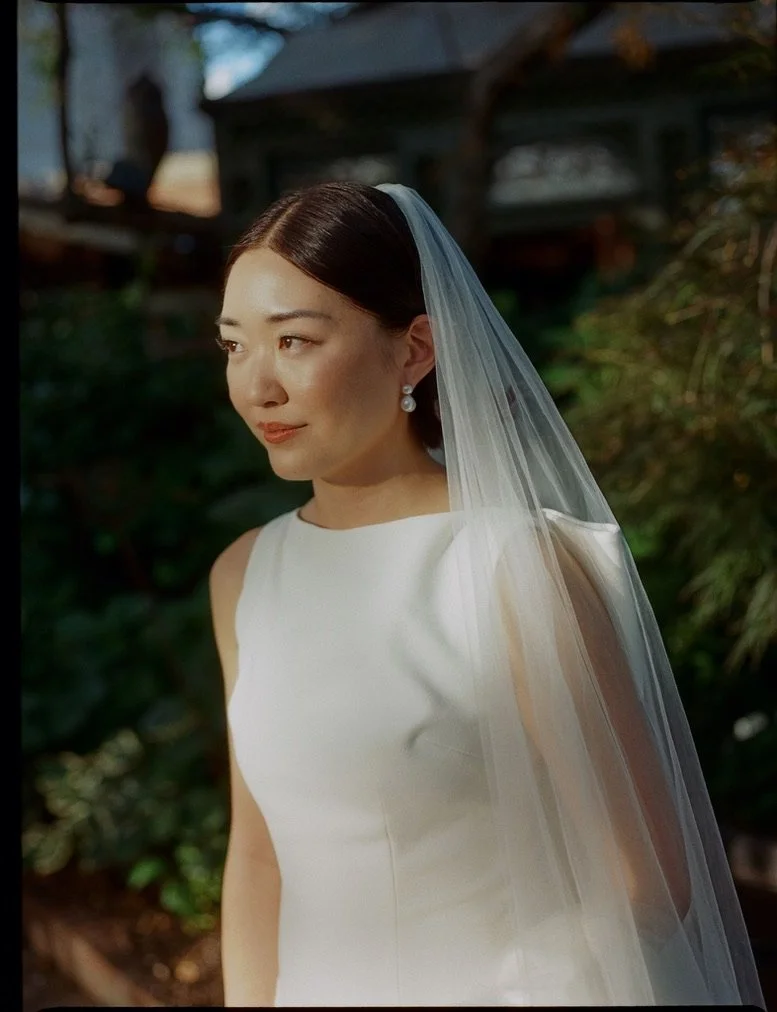 A woman dressed as a bride in a sleeveless white wedding gown with a sheer veil. She has dark hair styled neatly, wears pearl earrings, and is outdoors with green foliage and a house in the background, captured in soft lighting.