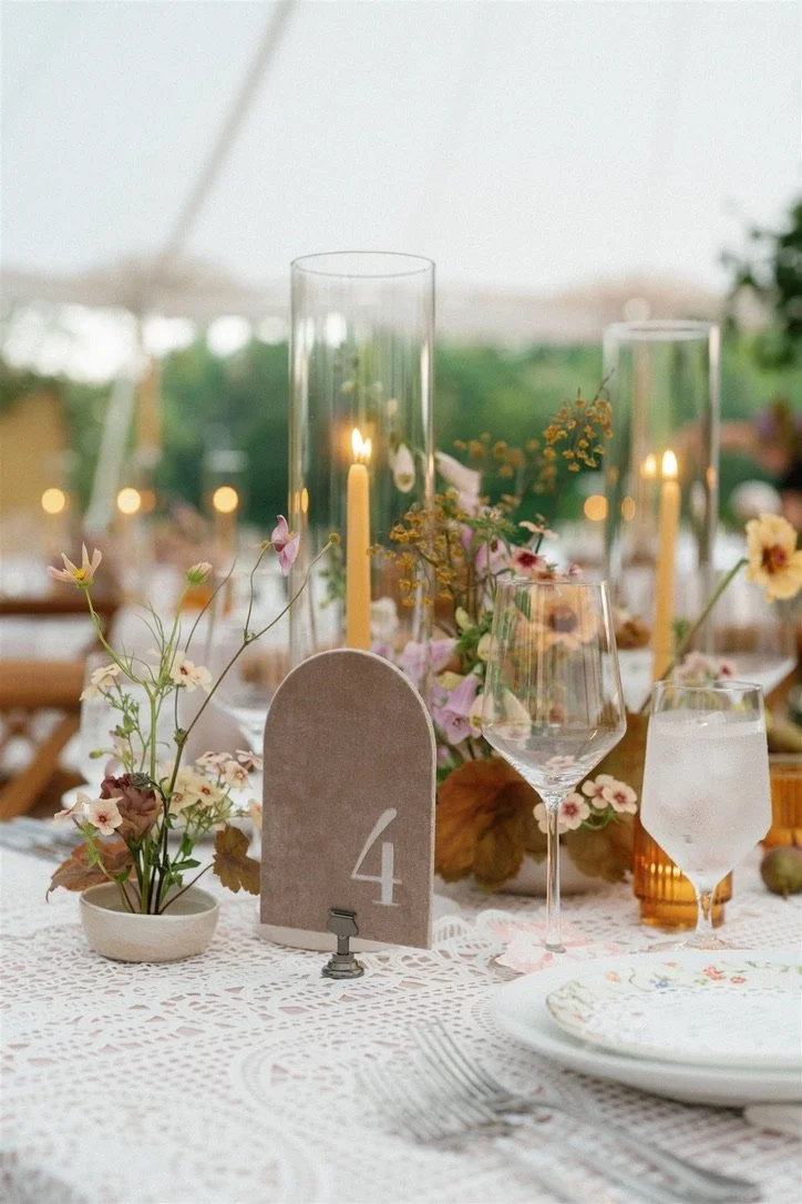 A decorated table with floral centerpieces, tall candles, glassware including wine and water glasses, a table number sign with the number 4, plates, and cutlery, set outdoors for a special occasion.