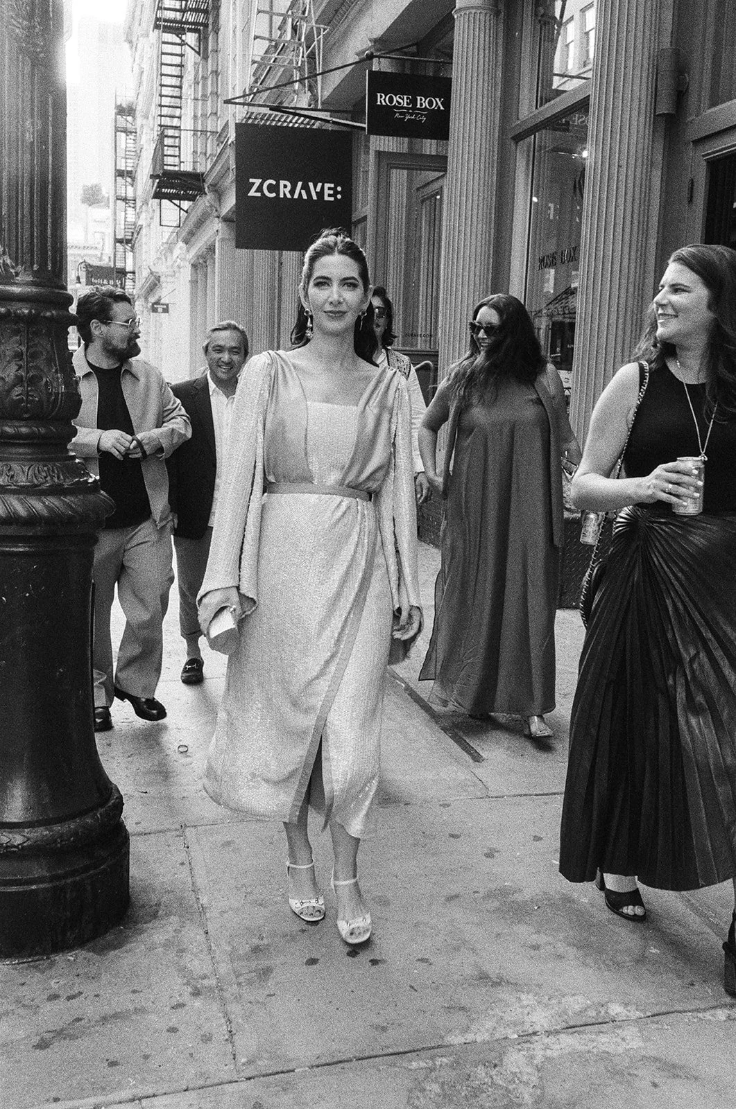 A group of women and men walking on a city sidewalk, with old buildings and signage including "ZCRAVE" and "ROSE BOX" in the background, black and white photograph.