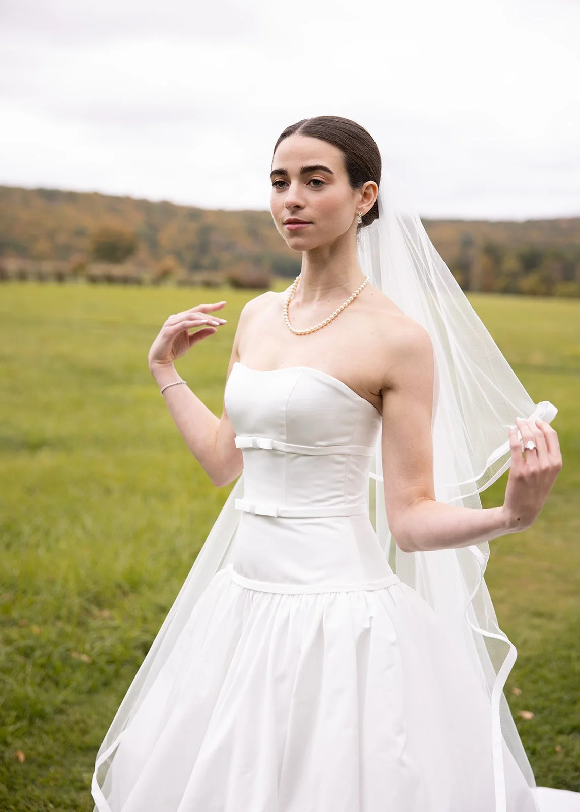 A bride in a white strapless wedding dress holding a veil outdoors in a rural landscape with green fields and hills in the background.