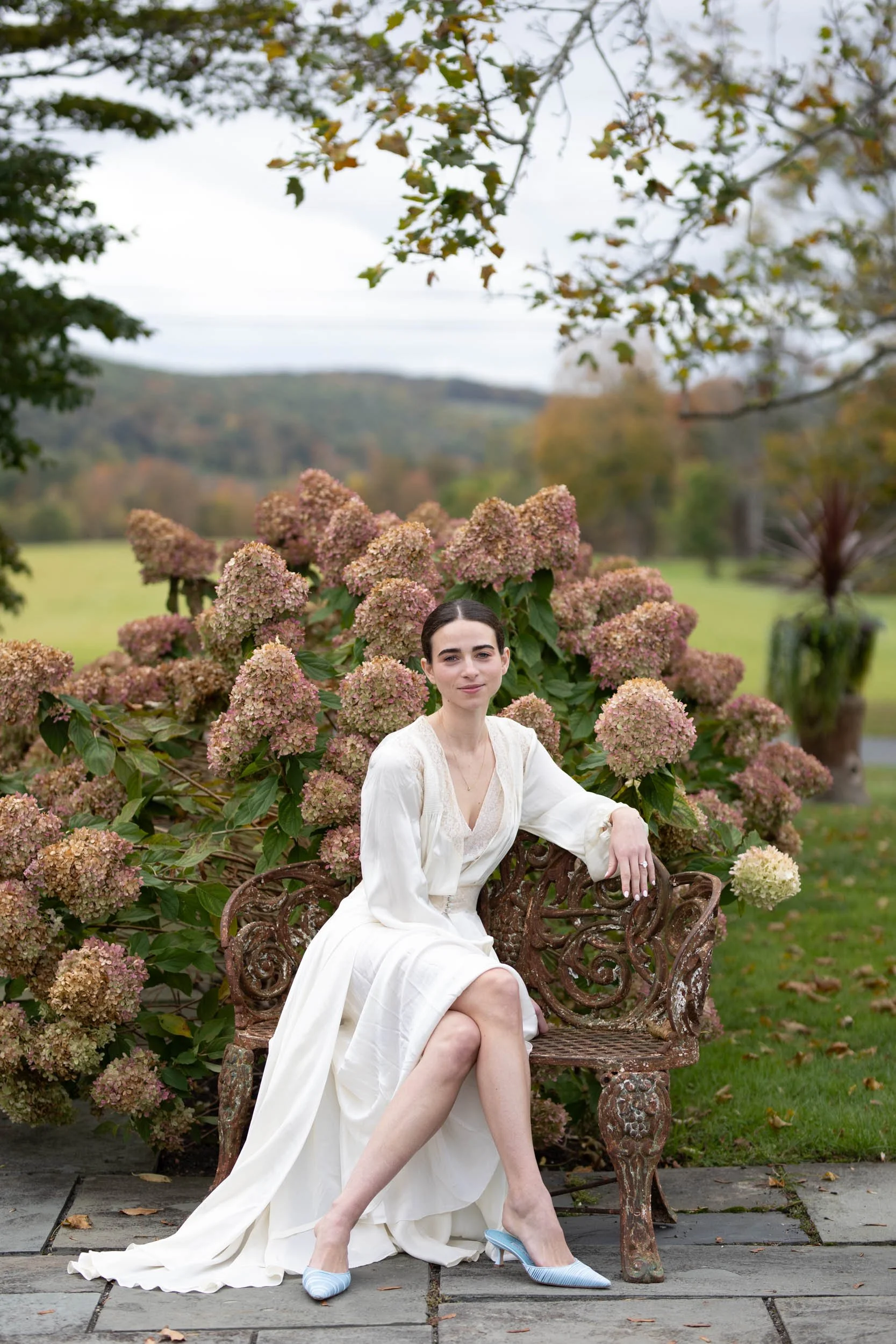 A woman in a white dress sitting on a decorative iron bench outdoors, surrounded by pink hydrangea flowers and trees with fall foliage, with mountains in the background.