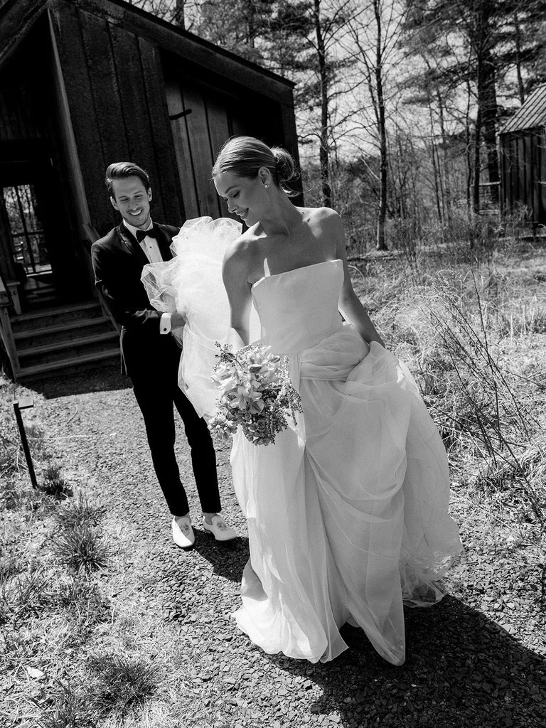 Black and white photo of a bride in a strapless wedding dress holding a bouquet, walking outdoors with a smiling groom in a tuxedo behind her, in a rustic setting with trees and wooden structures.