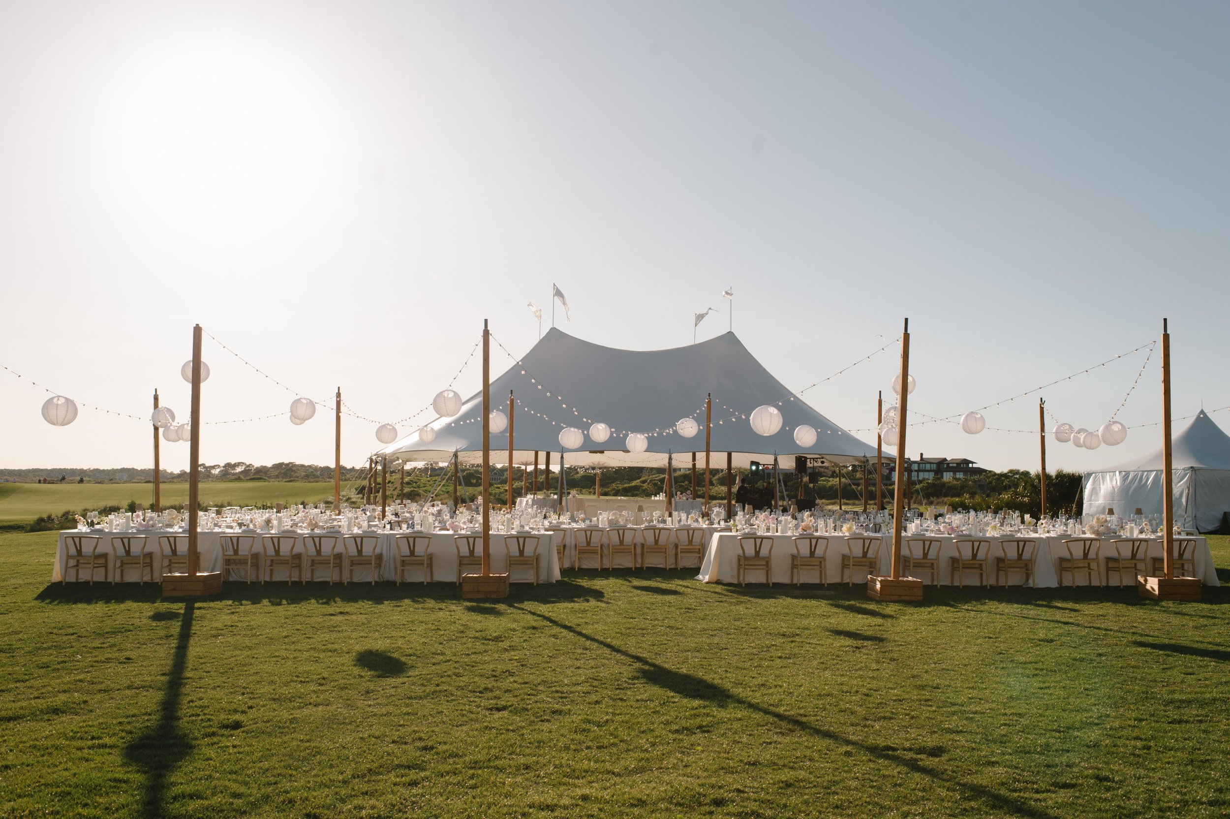 Outdoor wedding reception setup with long tables, wooden chairs, white tablecloths, and hanging lanterns under large white tent on grassy field during daylight