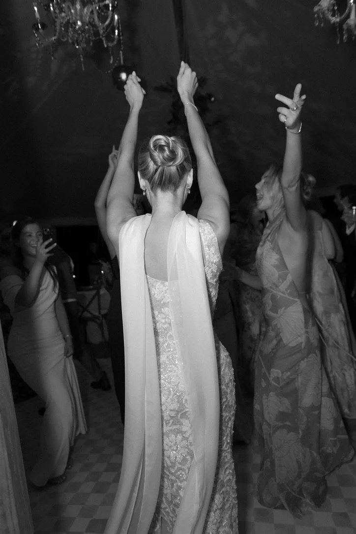 Women dancing at a celebration or party, with one woman in the center wearing a shiny dress and a chiffon shawl, raising her hands, and others around her smiling and enjoying.