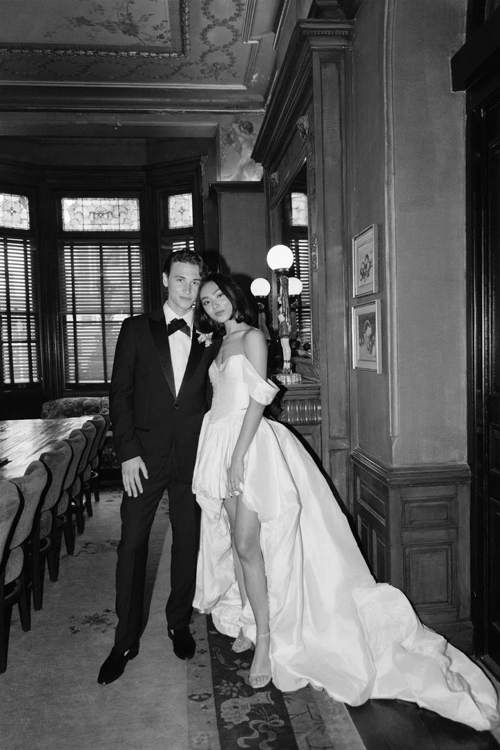 A black-and-white photo of a man and woman in formal attire inside a decorated room with large windows and wooden paneling. The man is wearing a tuxedo with a bow tie, and the woman is wearing a wedding dress. They are standing close together, posing