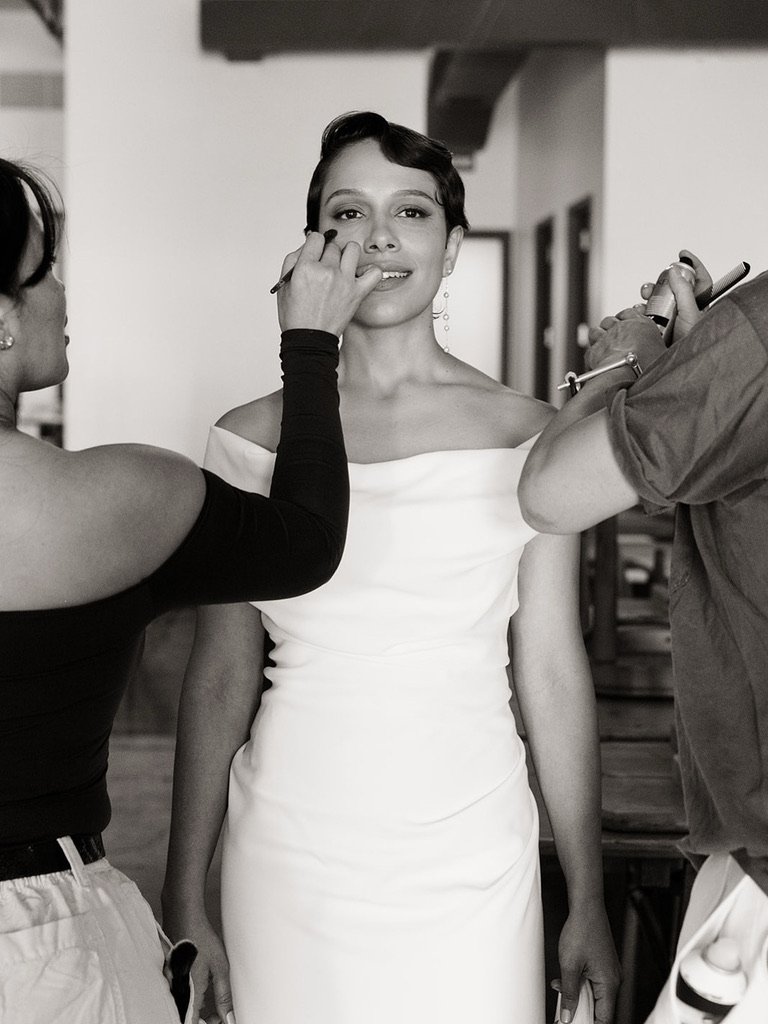 A woman getting her makeup touched up by a makeup artist while posing for a photo, wearing a white off-the-shoulder dress indoors.