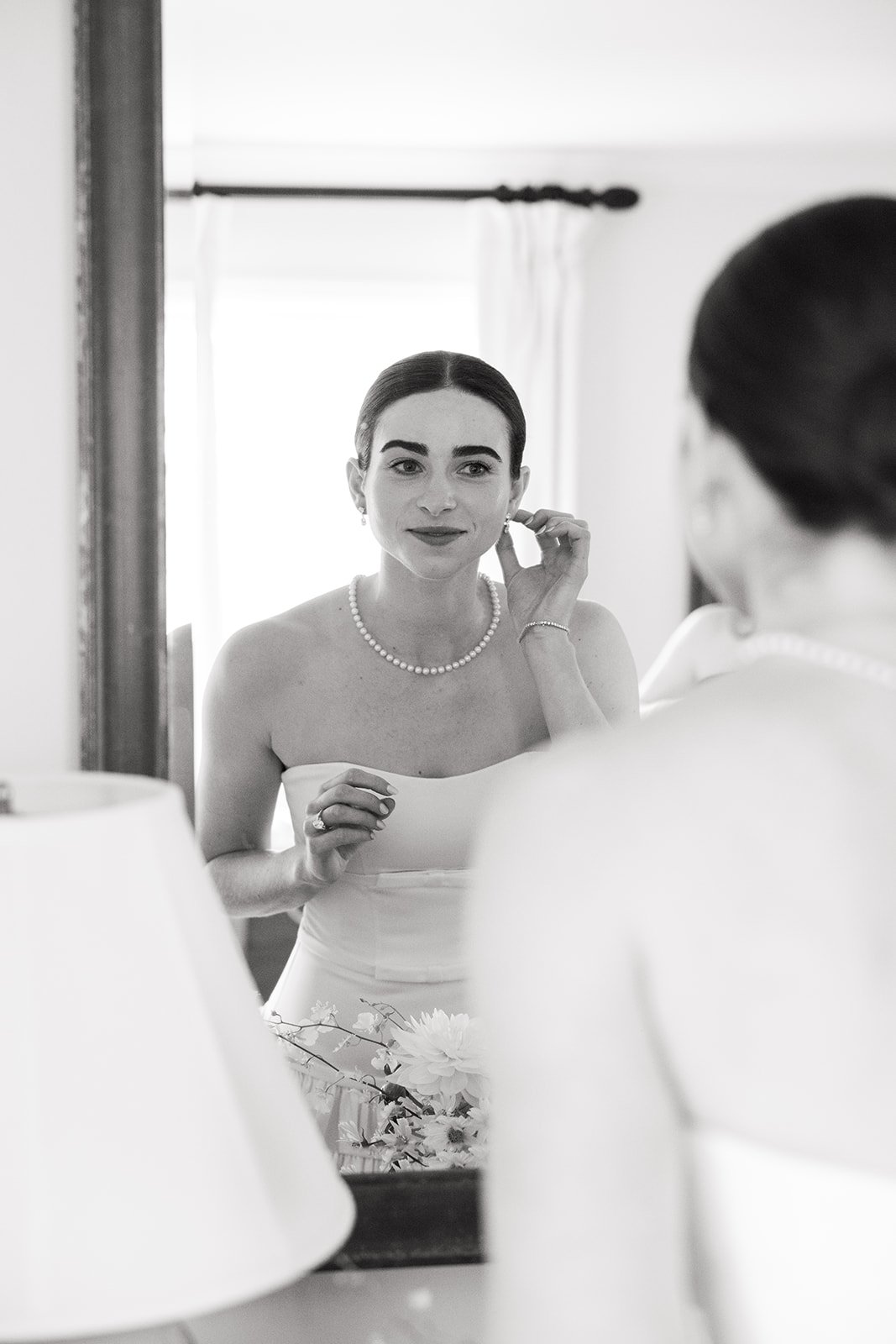 A woman in a strapless dress and pearl necklace looking at herself in a mirror, adjusting her earring.