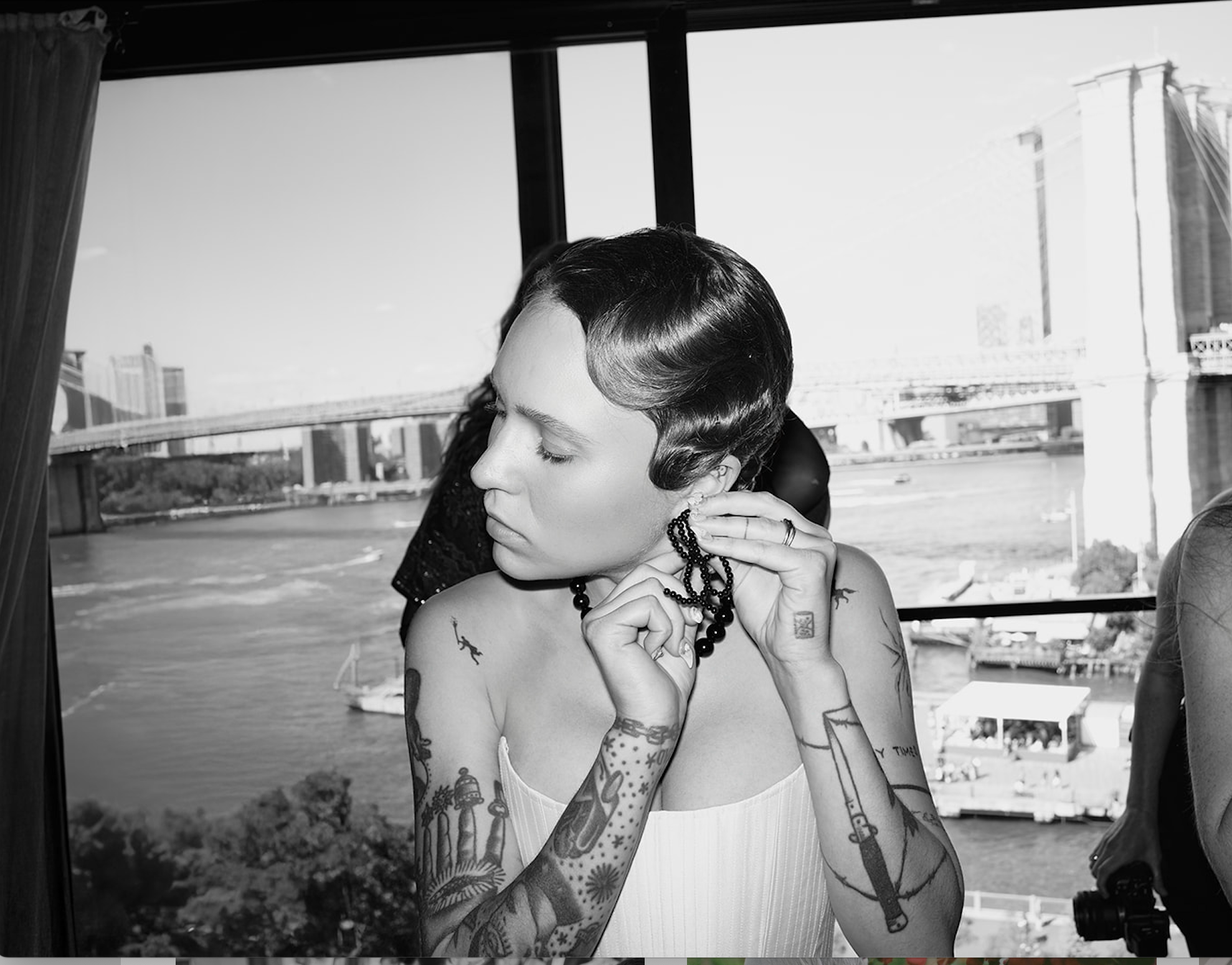 A woman with short dark hair and tattoos on her arms, wearing a white top, is putting on earrings in front of a window with a cityscape view, including a bridge and river in the background.