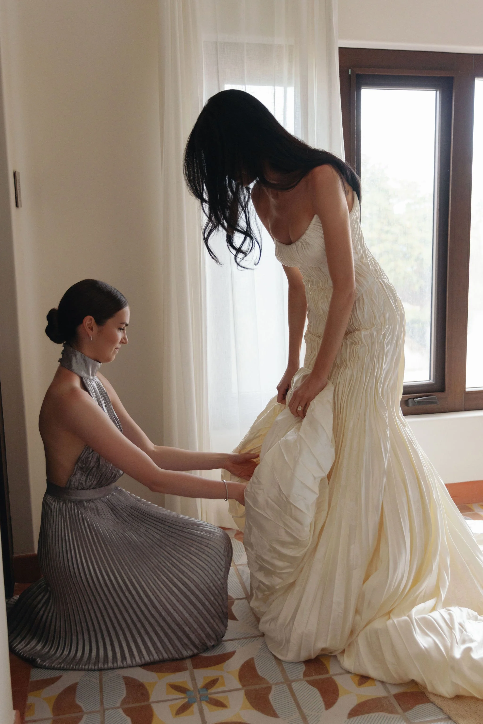 A woman in a wedding dress is being assisted with her gown by another woman in a gray dress, in a room with large windows and natural light.