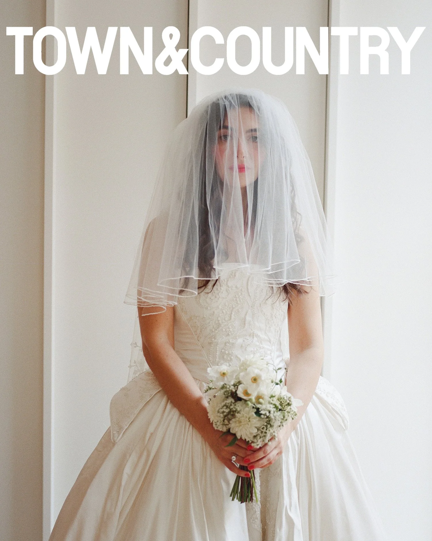 A bride in a wedding gown and veil holding a bouquet of white flowers, standing indoors with a neutral background.