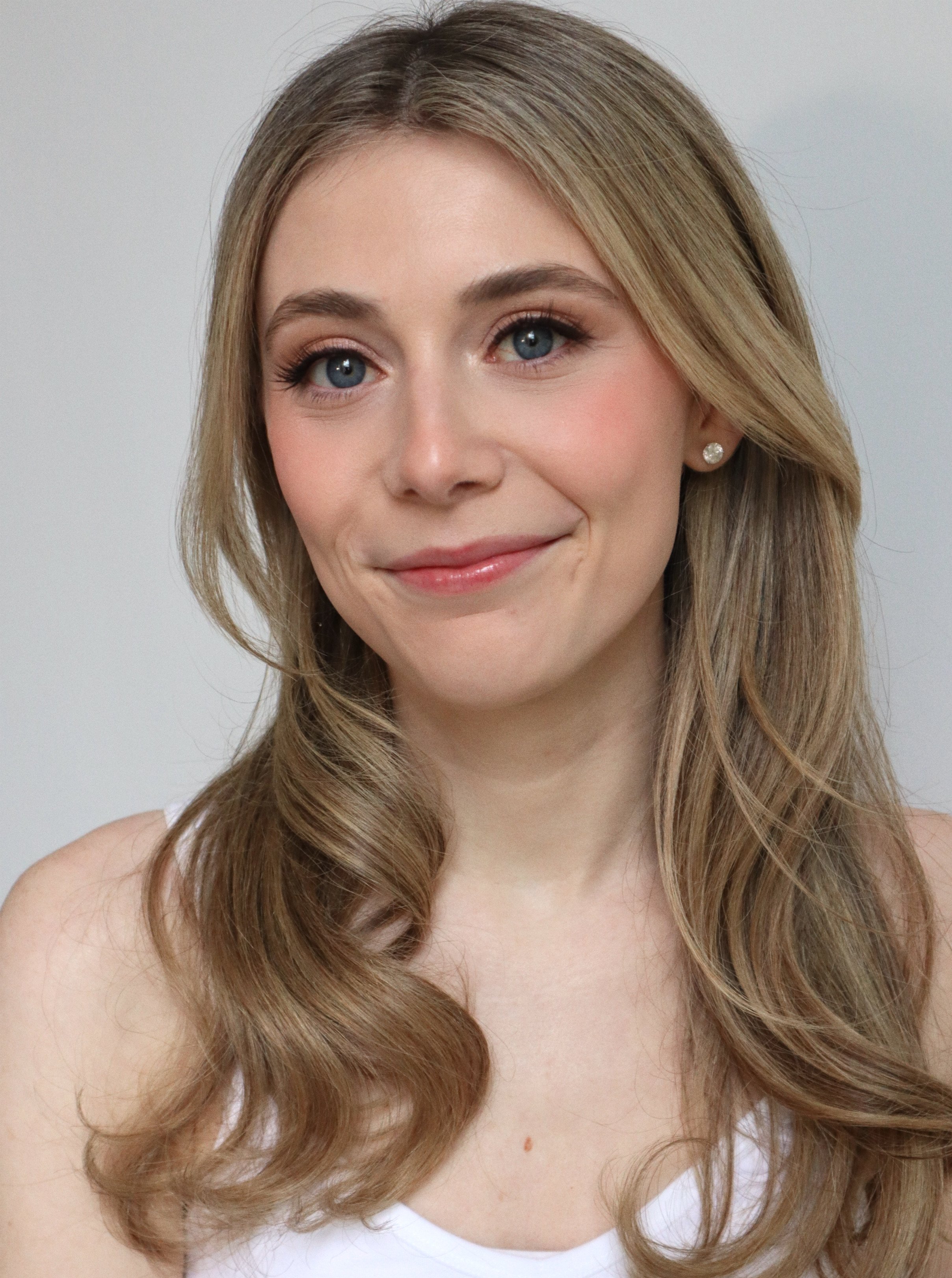 Close-up portrait of a young woman with blue eyes, long wavy light brown hair, and wearing pearl earrings, smiling softly against a plain background.