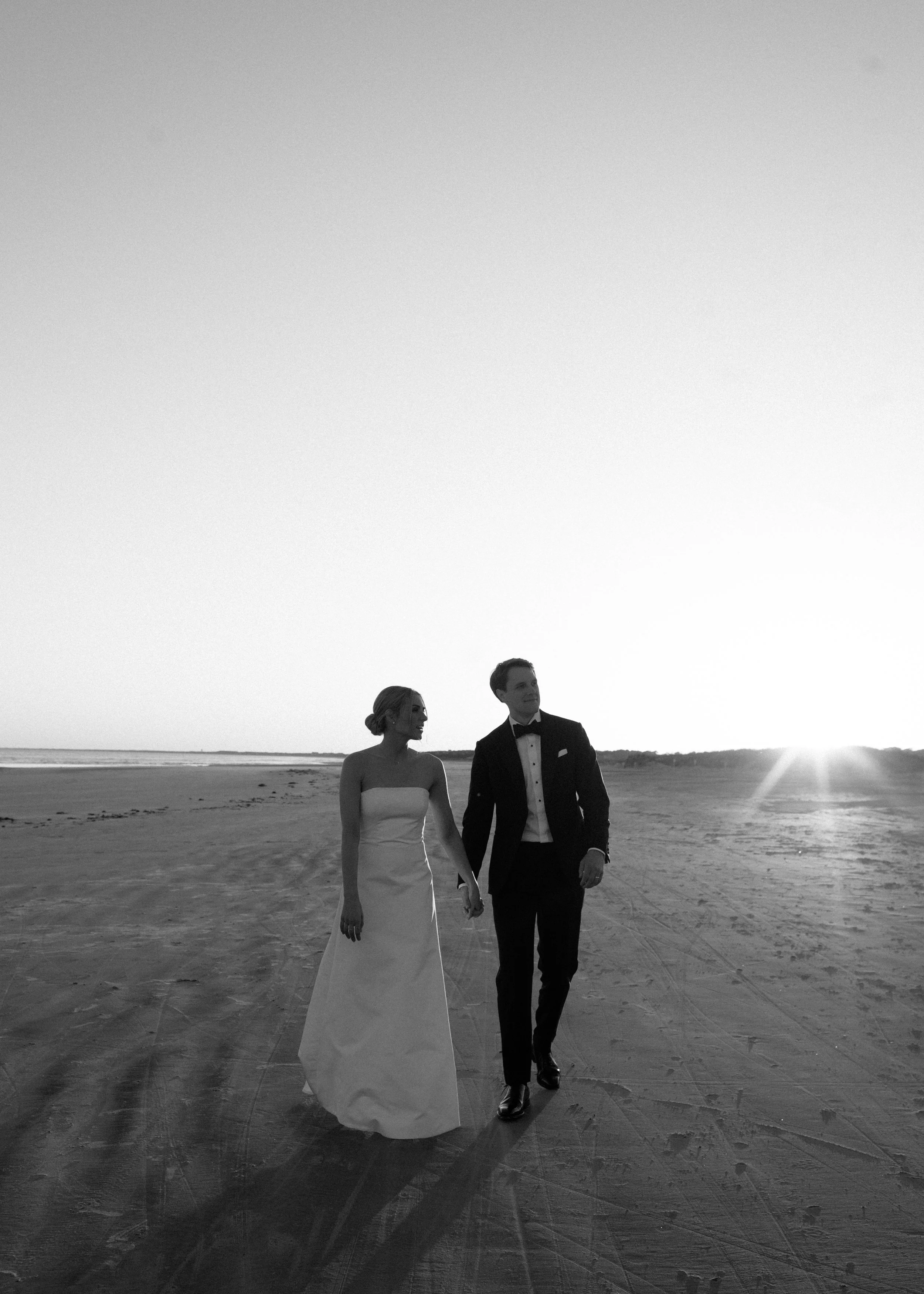 A black and white photo of a bride and groom walking hand in hand on a beach at sunset.
