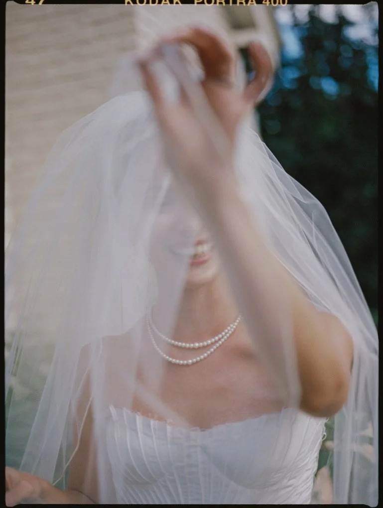 A bride in a white wedding dress and pearl necklace, smiling behind a veil, outdoors with a blurred background.