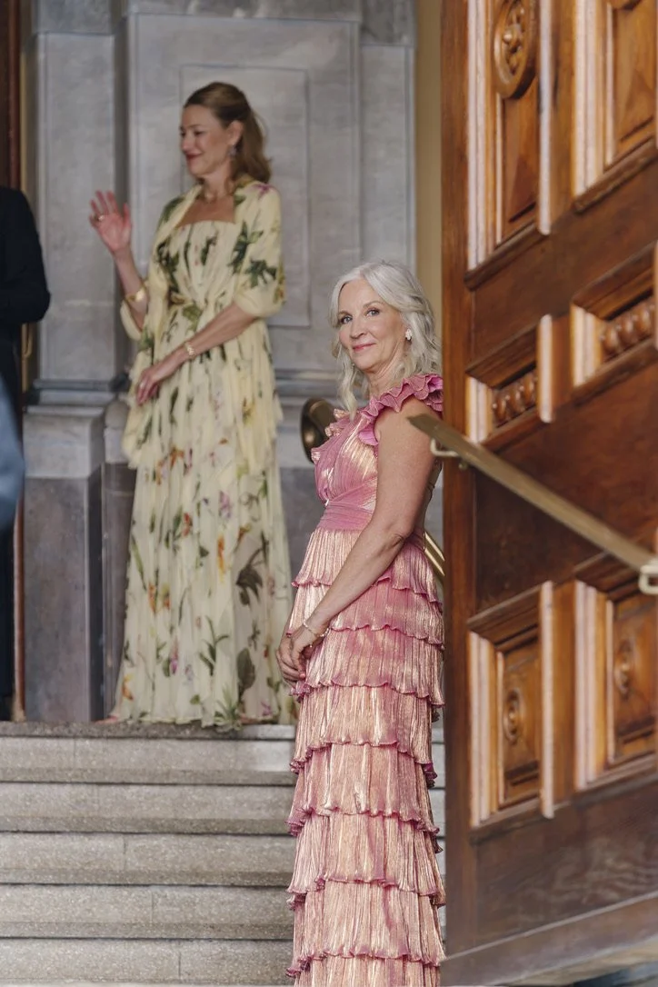 A woman with gray hair in a pink ruffled dress standing on a staircase, smiling and looking to the side. In the background, another woman with brown hair in a floral dress is visible.