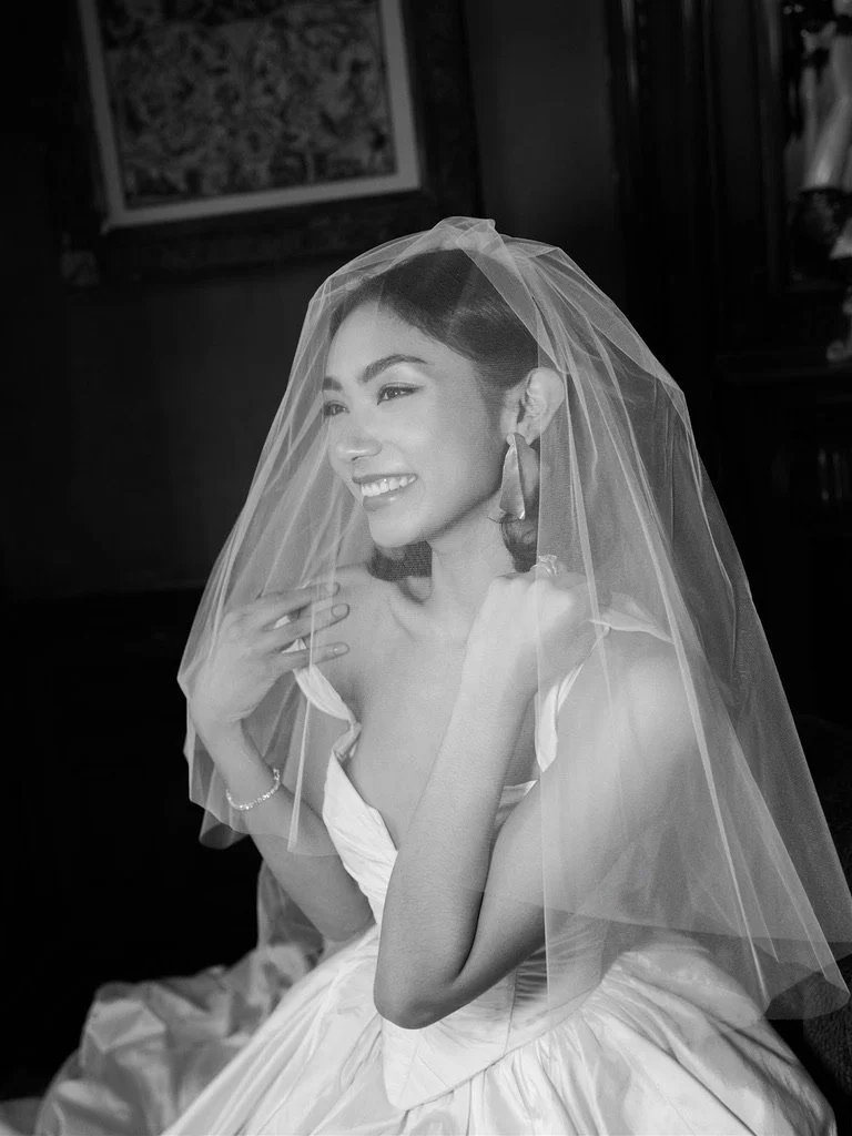 A smiling woman in a wedding dress with a veil, sitting indoors in black and white.