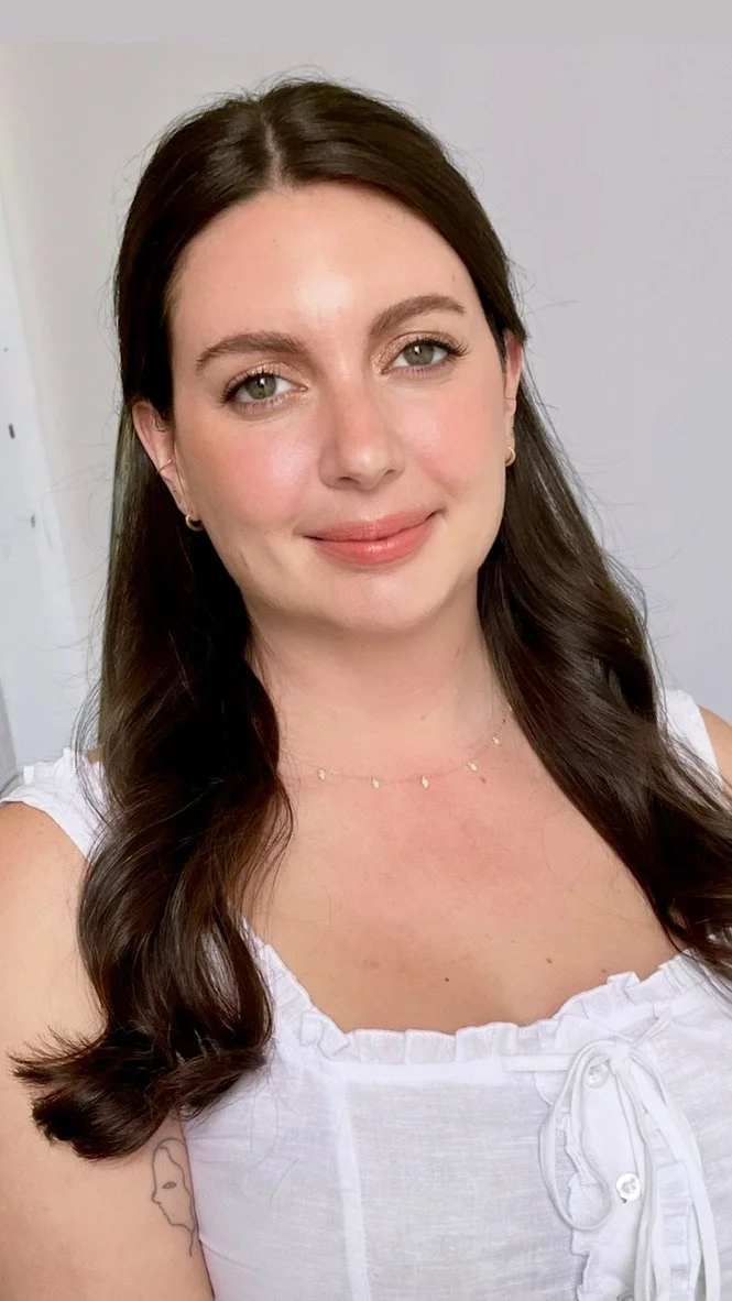 A young woman with long dark brown hair, light skin, and green eyes, smiling slightly, wearing a white sleeveless top with ruffle details and a delicate gold necklace, standing indoors against a neutral gray background.