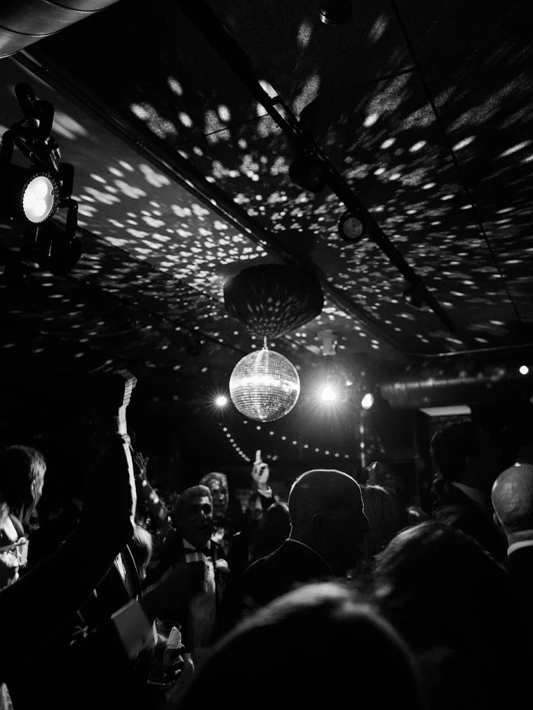 People dancing at a party under disco ball with light reflections on ceiling and walls.