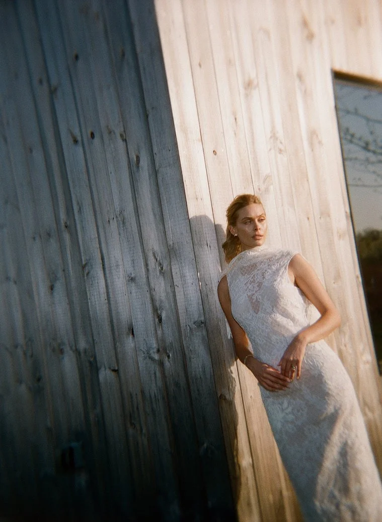 A woman in a white lace dress leaning against a wooden wall outdoors at sunset.