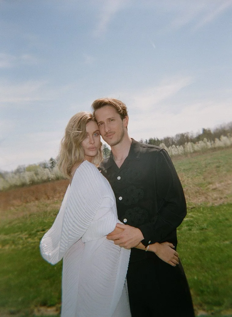 A young couple standing close together outdoors, with the woman in a white dress and the man in a black jacket, under a partly cloudy sky, on a grassy area with trees in the background.
