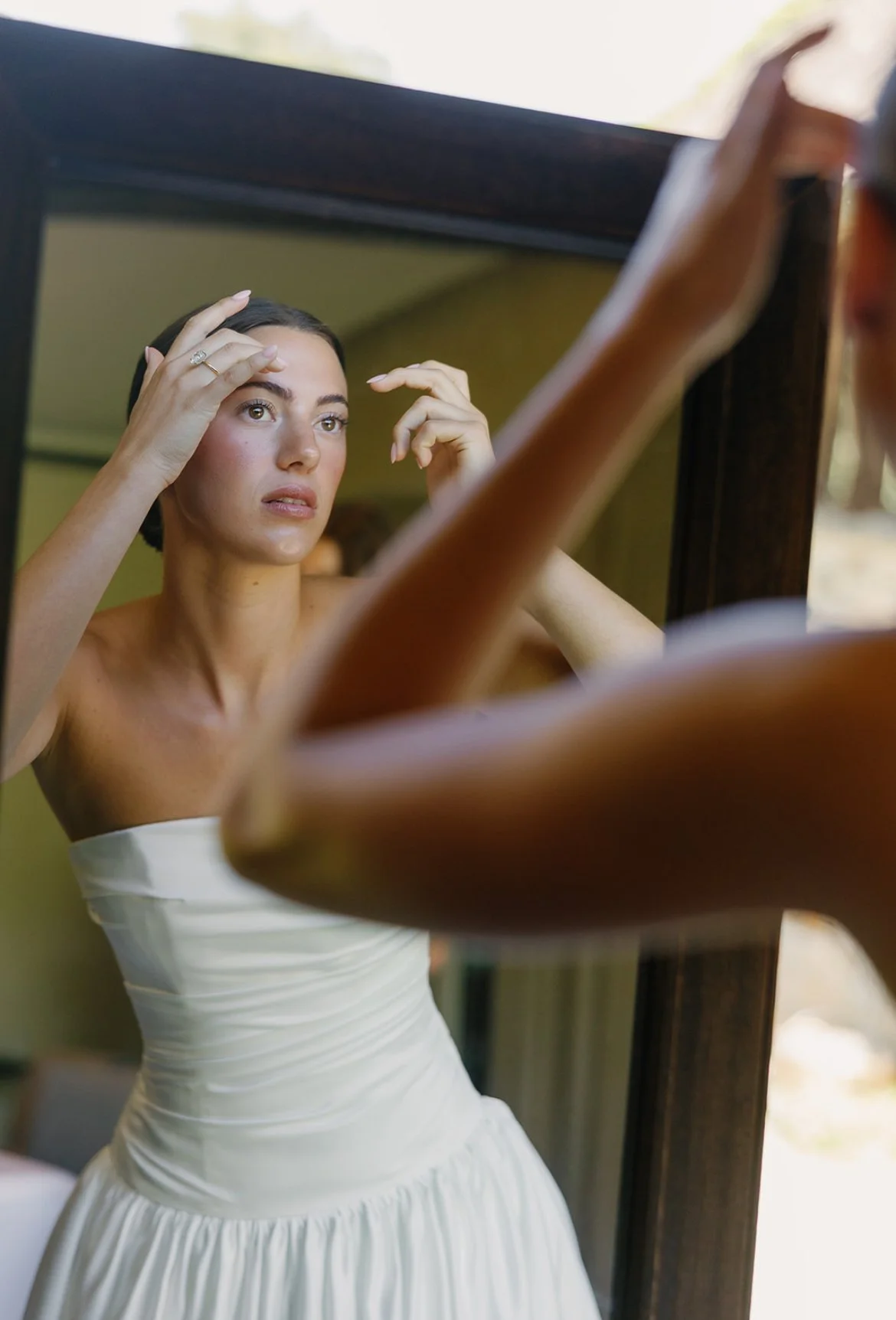 A woman in a white strapless dress looking at herself in a mirror, adjusting her hair, with an outdoor background visible through the window.