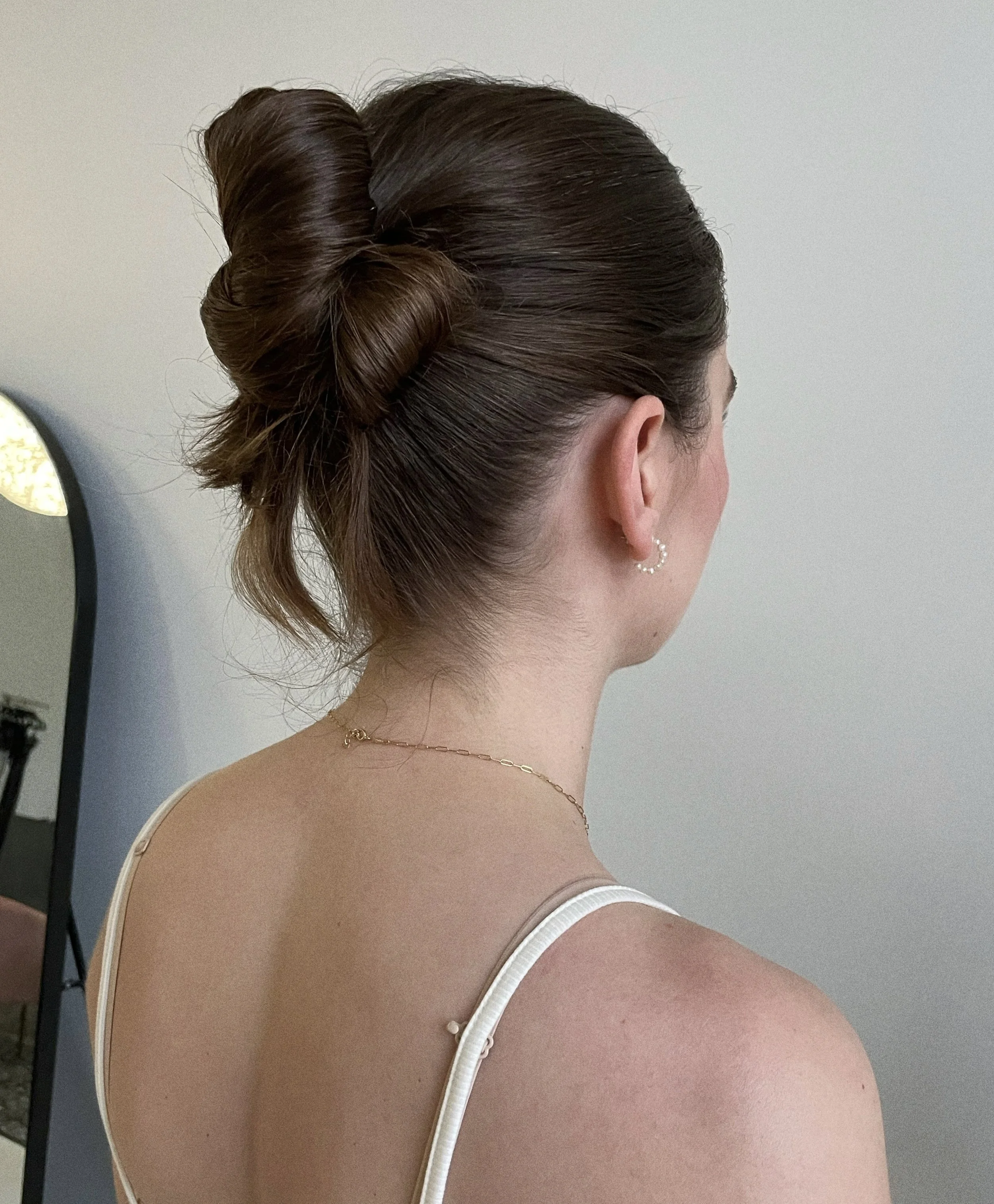 Back view of a woman with styled brown hair in an updo and wearing a white spaghetti strap top, small earrings, and a delicate necklace.