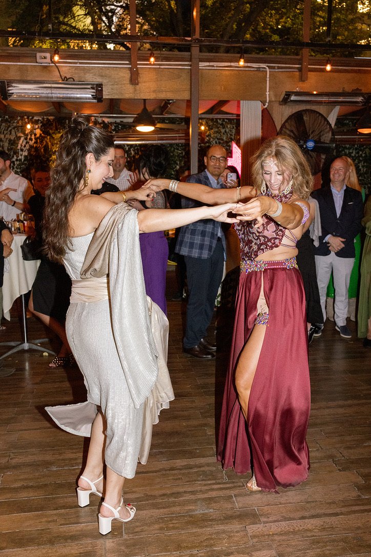 Two women dancing together at a party or celebration, holding hands and smiling. One woman is wearing a white dress with a beige shawl, and the other is in a burgundy outfit with a slit skirt. People can be seen watching and enjoying in the backgroun