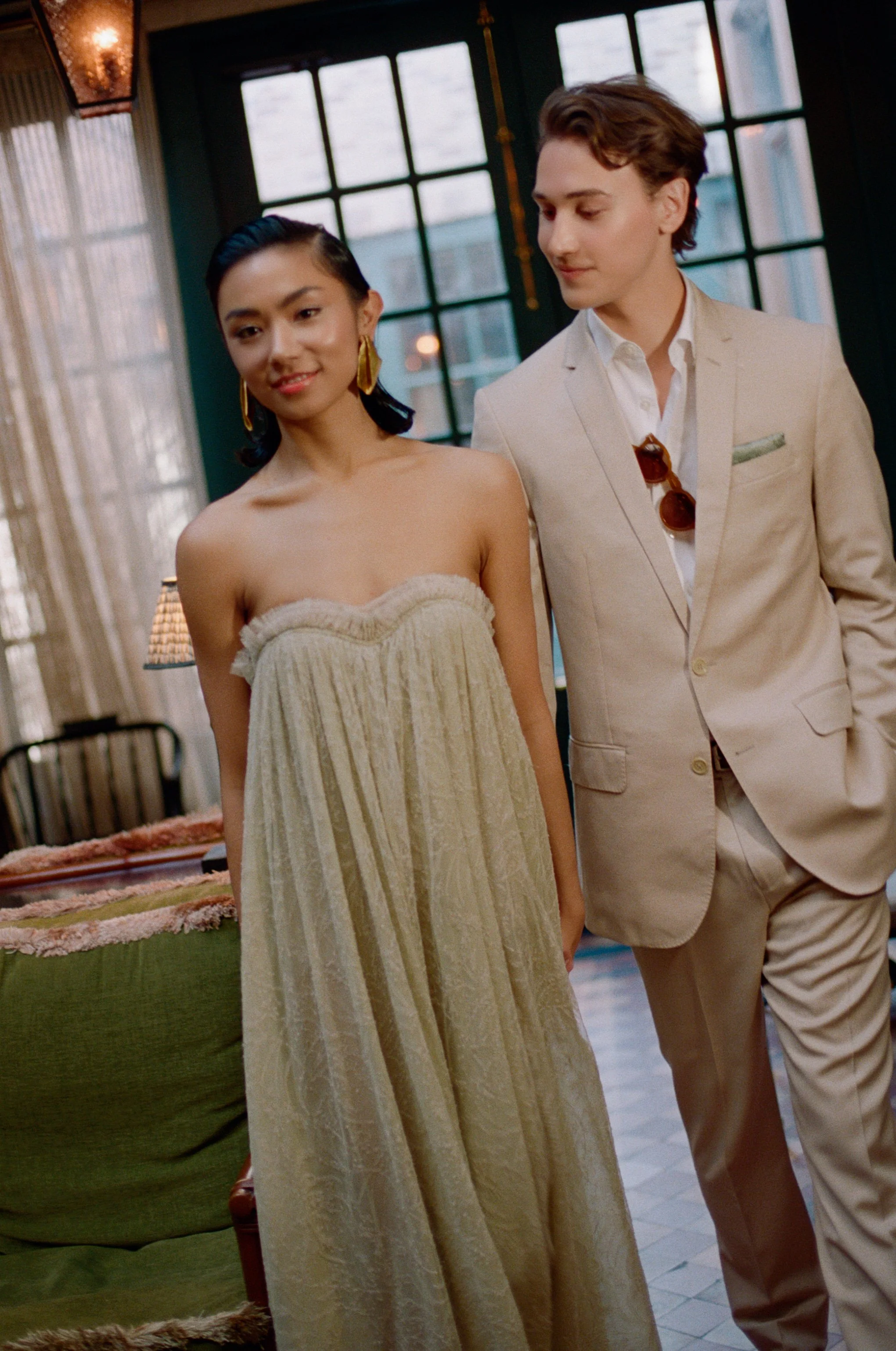 A woman in a strapless beige dress and gold earrings standing next to a man in a cream-colored suit inside a well-lit room with large windows.