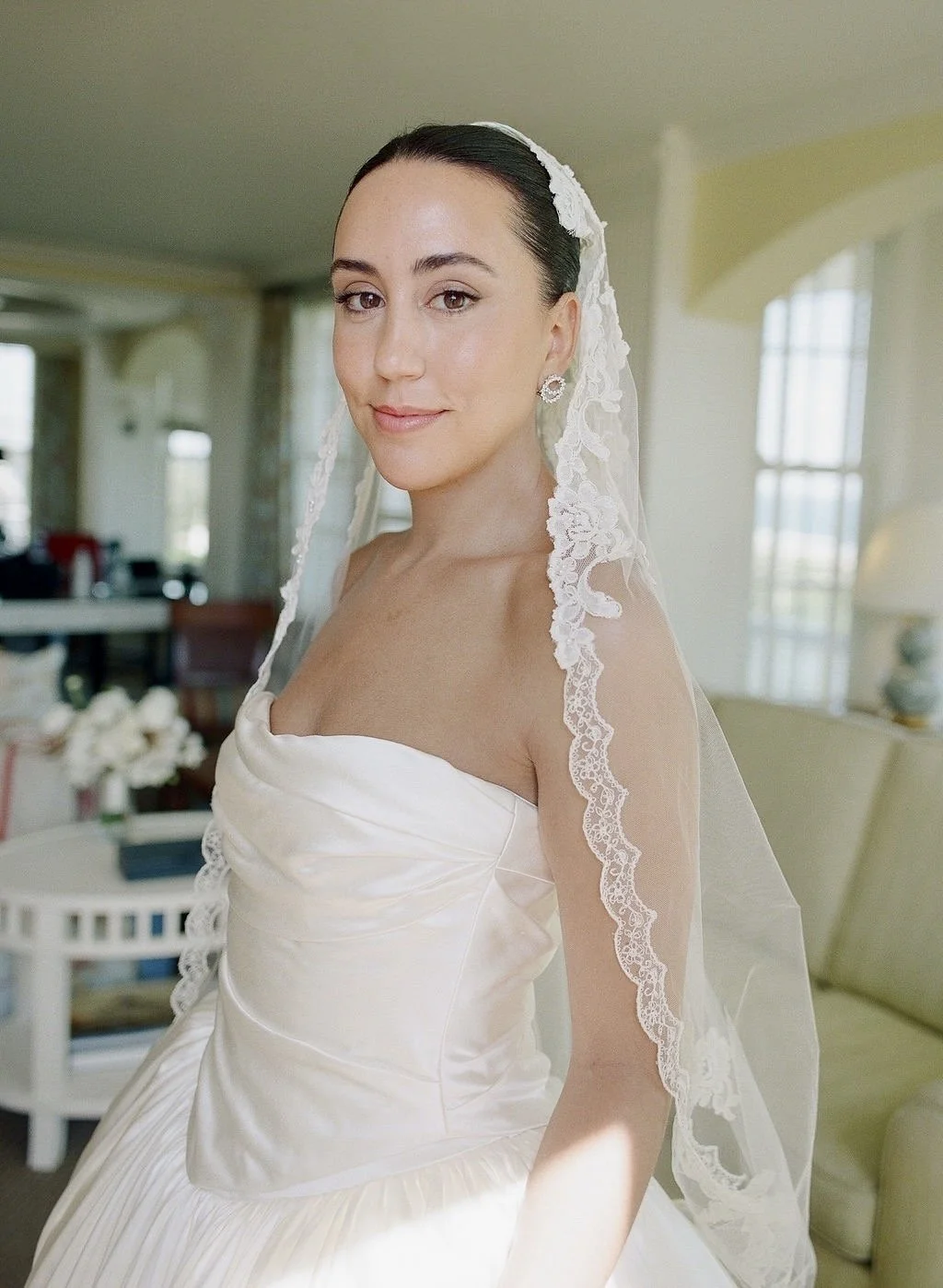 Close-up of a bride in a satin wedding dress with lace veil, standing in a well-lit room with large windows and yellow walls.