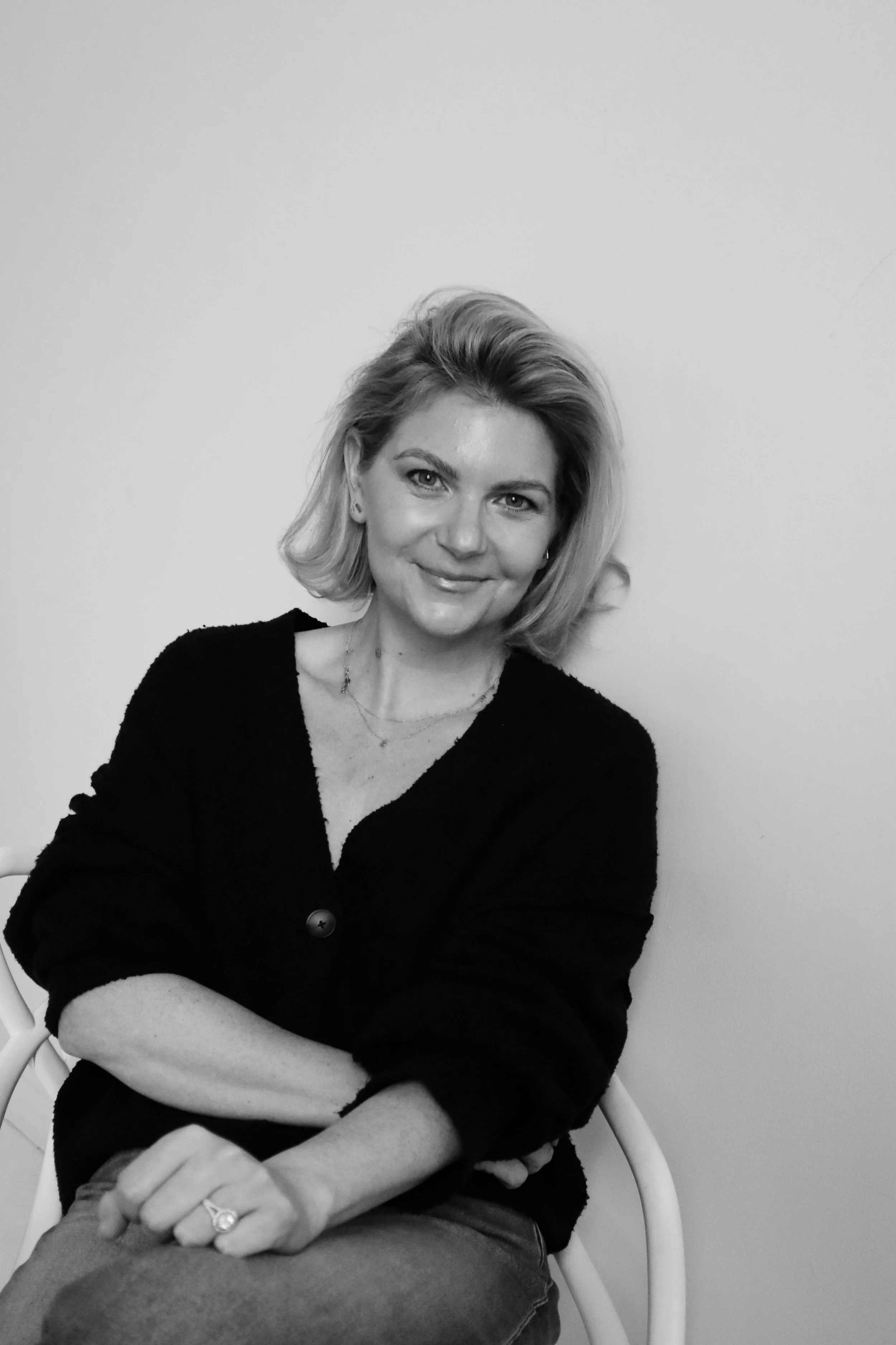 A woman with blonde hair smiling and sitting on a white chair, wearing a black sweater and textured necklace, against a plain white wall.