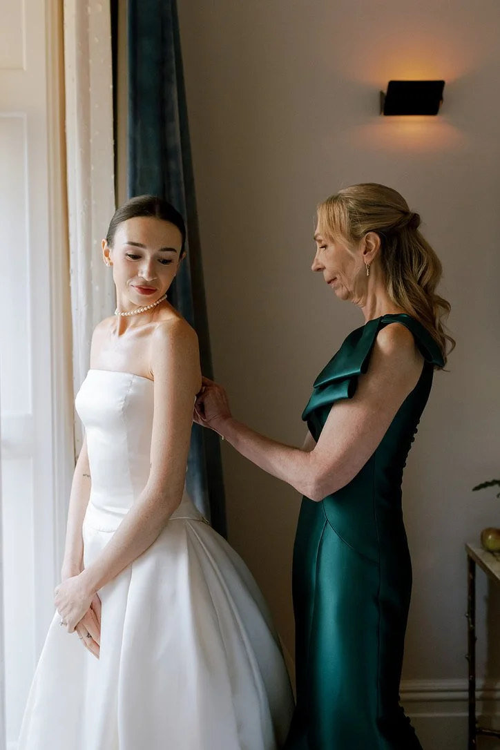 A bride in a white wedding gown standing by a window, with an older woman, likely her mother, helping her adjust her dress in a warmly lit room.