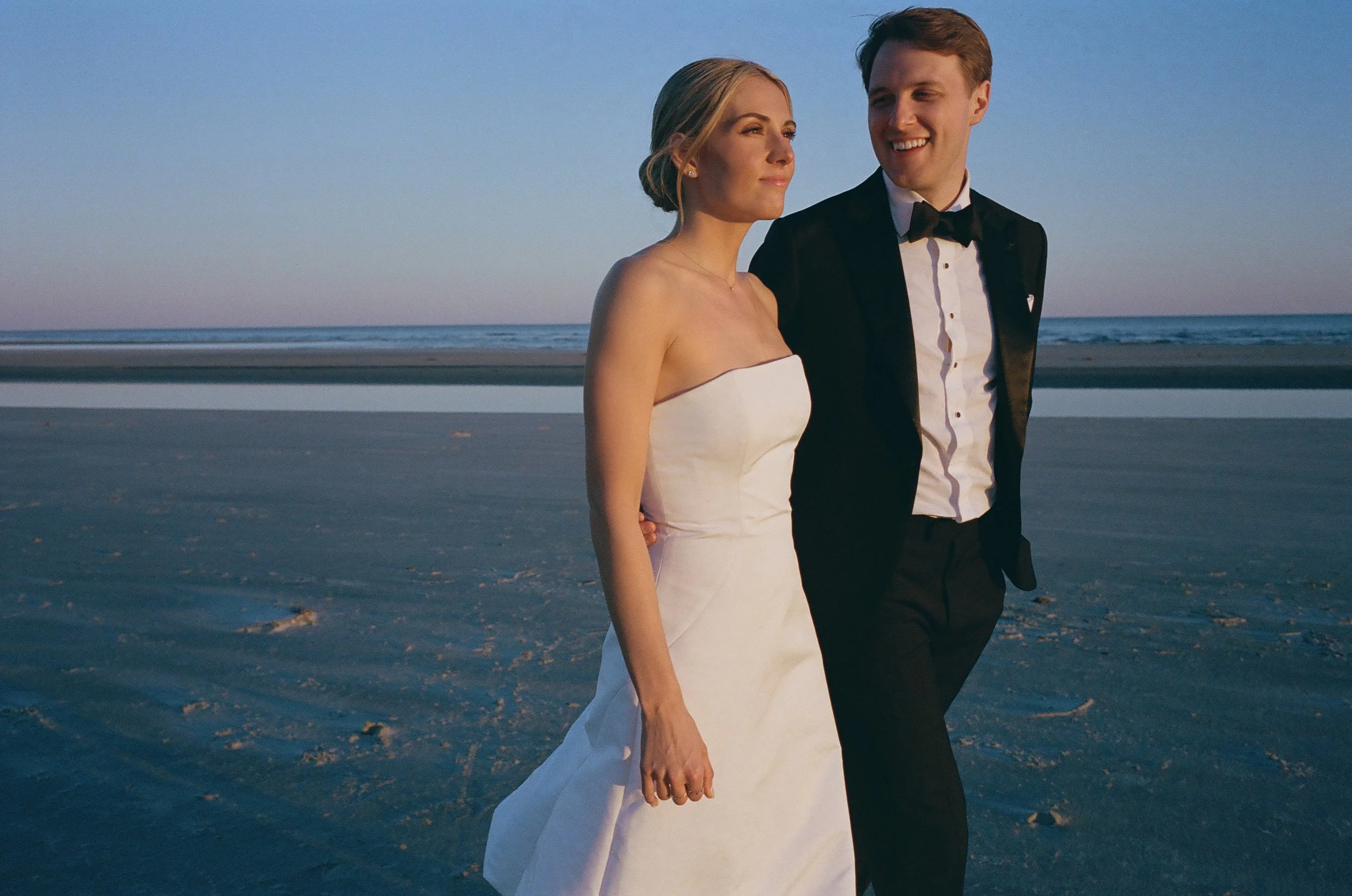 A newlywed couple, a woman in a white strapless wedding dress and a man in a black tuxedo, walking on the beach during sunset or twilight.