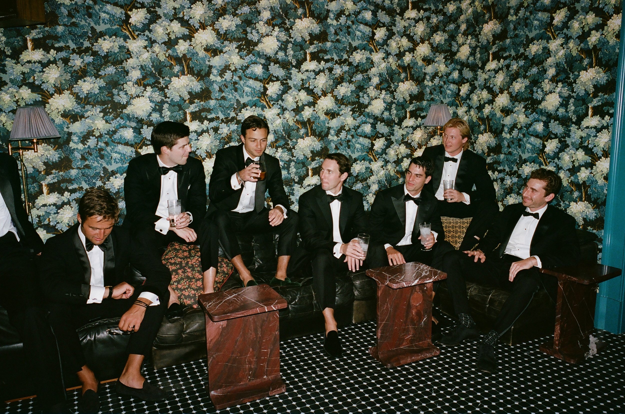 Seven men in tuxedos sitting and standing in a room with floral wallpaper, drinking beverages and socializing.