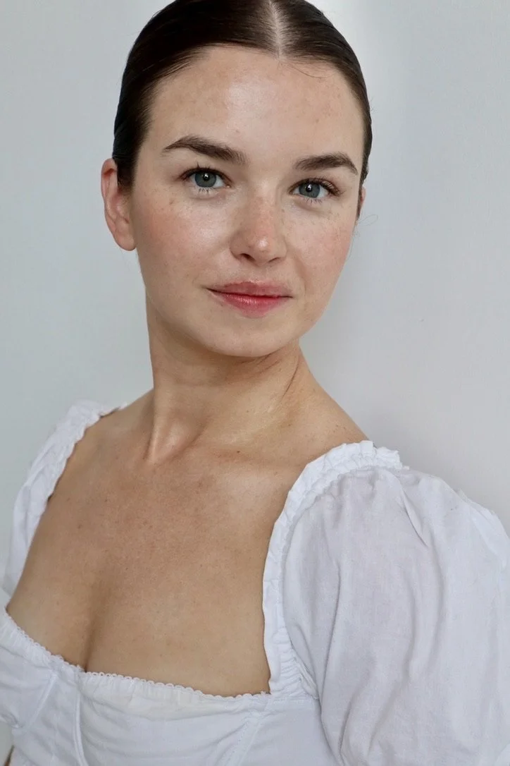 Portrait of a young woman with fair skin, blue eyes, and brown hair pulled back, wearing a white top with puffed sleeves, standing against a plain light gray background.