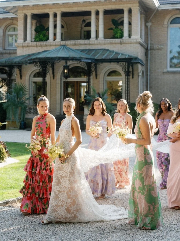 Group of women in colorful dresses standing outside a large brick mansion, holding bouquets, with one woman in a white wedding gown at the center.
