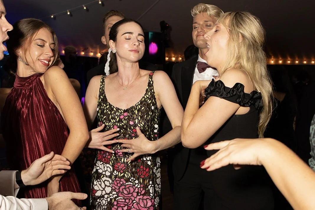 Group of people at a party, some appearing to be in a playful or flirtatious situation, with one woman holding her hands on her stomach, surrounded by smiling and amused individuals.