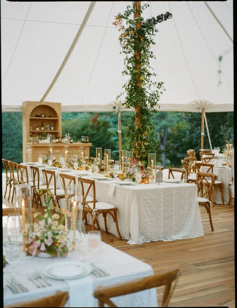 Elegant outdoor wedding reception setup inside a large white tent with long tables covered in white lace tablecloths, wooden chairs, and floral centerpieces. A tall wooden pole with greenery and floral decorations divides the space, with a rustic woo