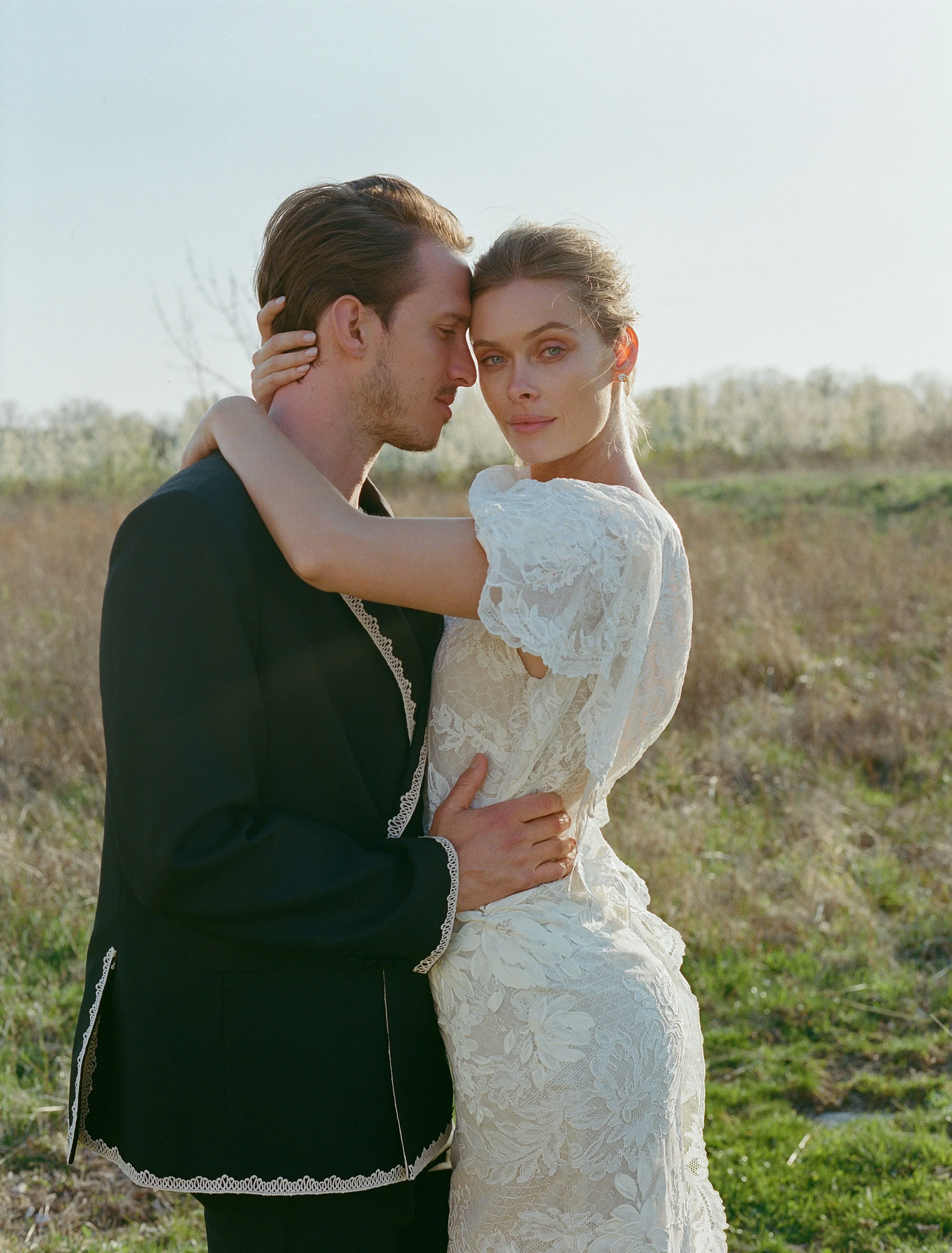A couple dressed in wedding attire standing close together outdoors, with the woman’s arms around the man's neck and the man holding her waist, in a grassy field with a clear sky.