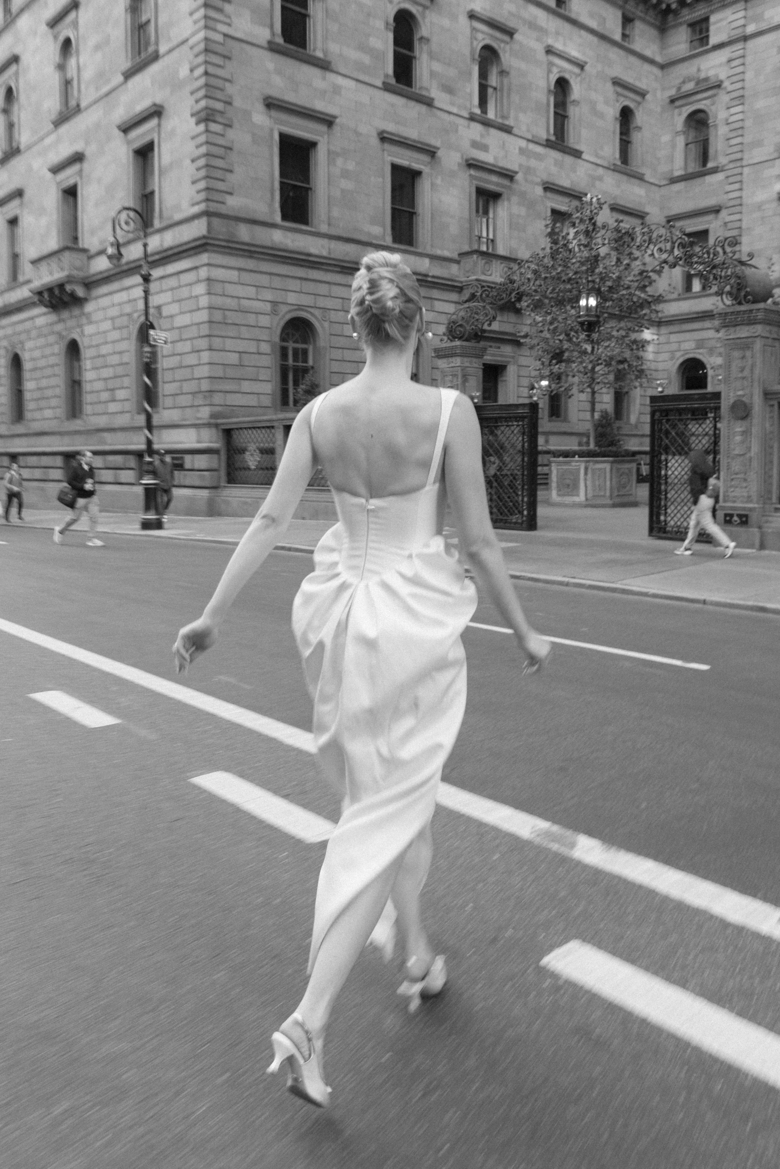 A woman in a white dress and high heels crossing a city street, with classical stone buildings and people walking nearby.