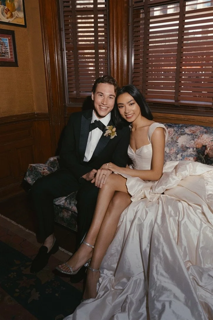 A young couple dressed in formal attire sitting together on a floral sofa in a room with wooden paneling and window shutters, smiling at the camera.