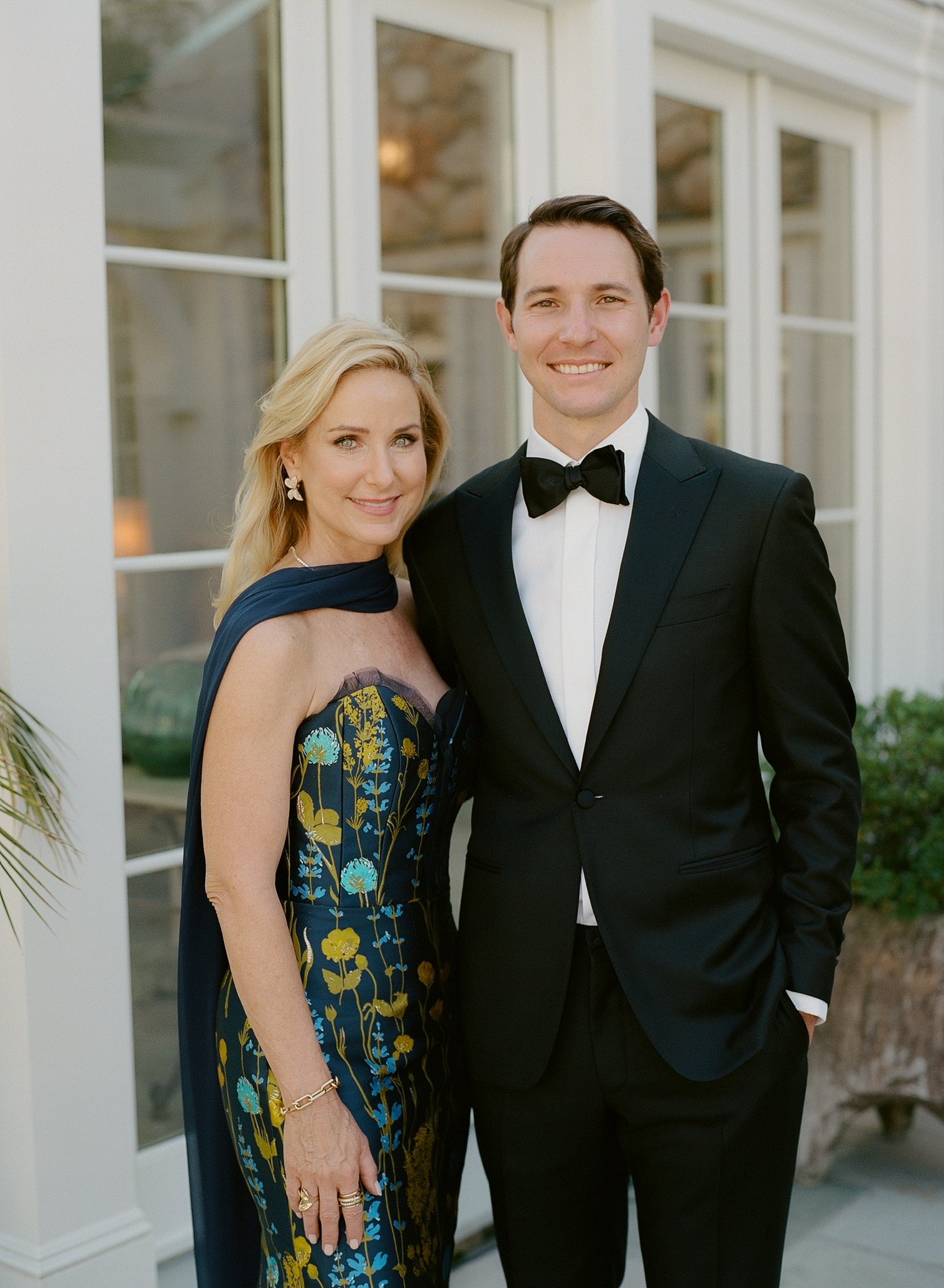 A woman in a strapless floral dress with a dark blue shawl and a man in a black tuxedo with a bowtie, standing together, smiling at a formal event.