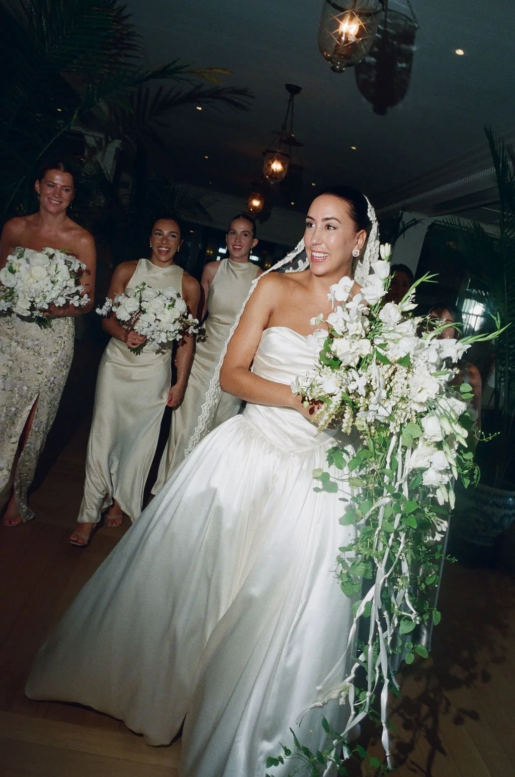 A bride in a white wedding dress holding a large bouquet of white flowers, standing at a wedding altar decorated with white flowers and greenery. Four bridesmaids, all in cream-colored dresses and holding smaller bouquets, are smiling and standing be