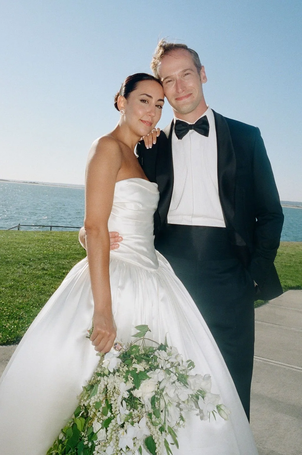 A bride and groom in wedding attire standing outdoors by the water, with the bride holding a large floral bouquet.