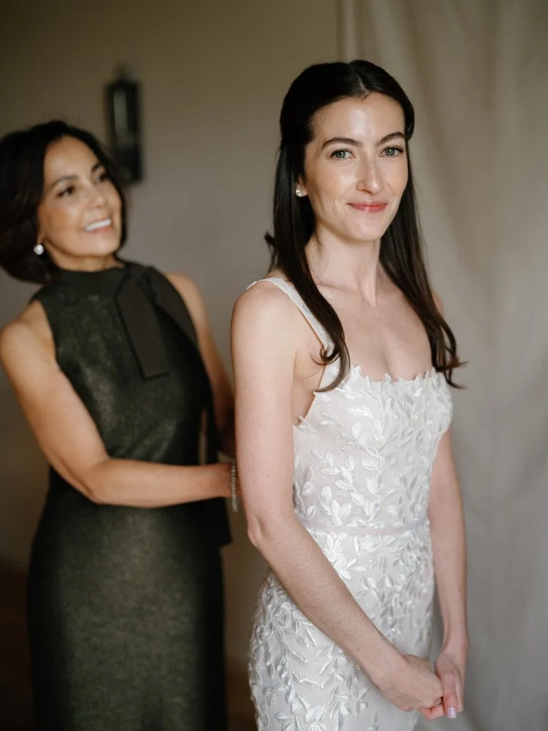 A woman in a white, floral wedding dress with dark hair is smiling softly at the camera. Behind her, another woman in a dark dress is smiling and looking at her.