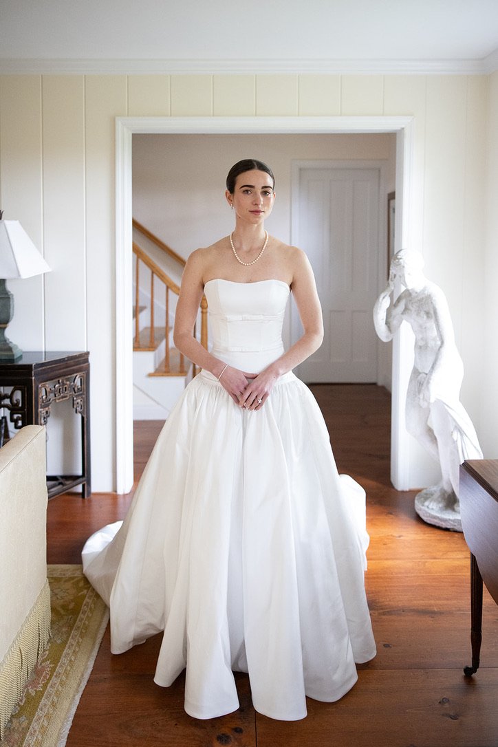 A woman in a strapless white wedding dress with a full skirt inside a home, standing in front of a doorway, wearing pearl necklace and bracelet, with a sculpture of a woman on her right side and a staircase in the background.