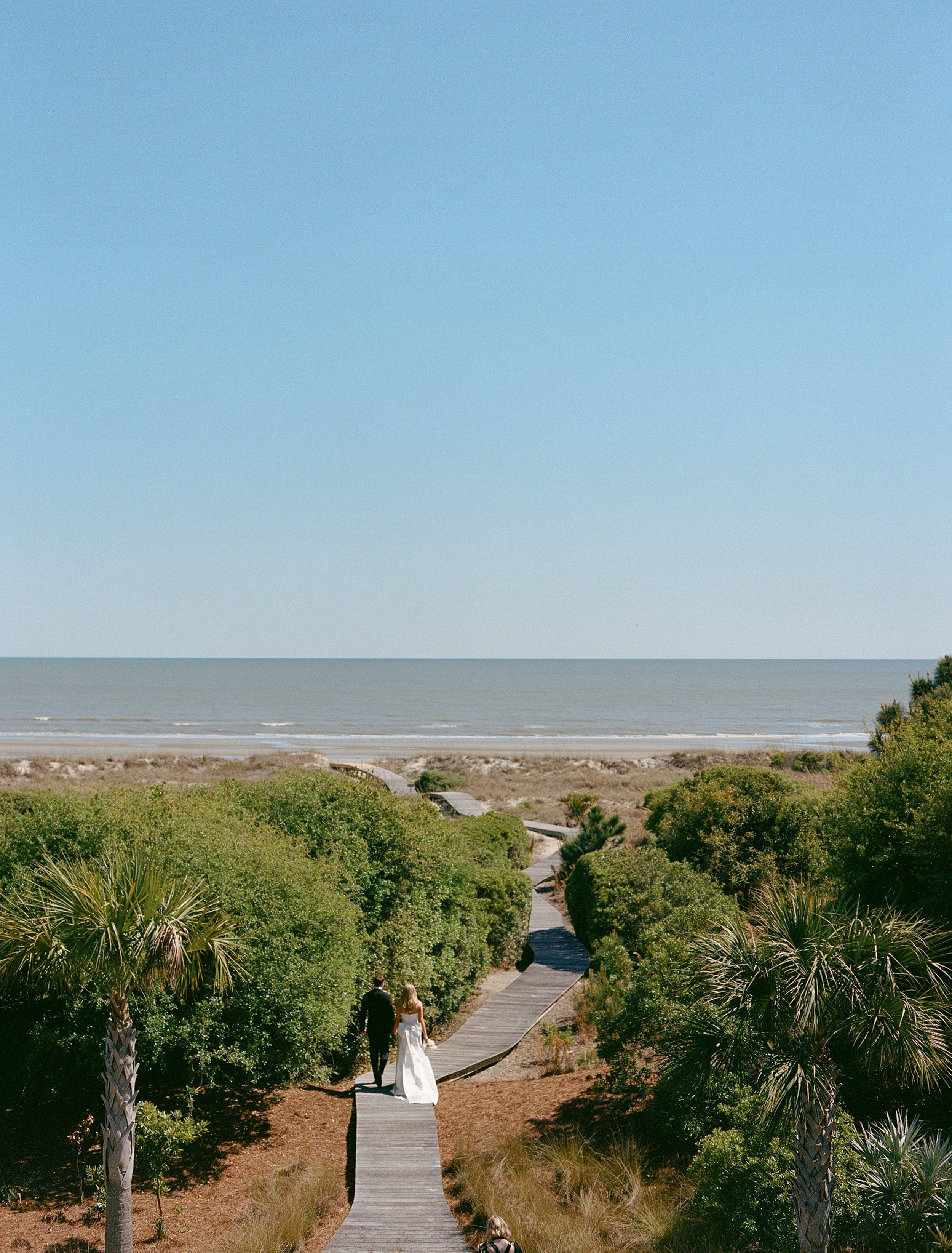 A couple in wedding attire walking hand in hand on a wooden pathway through lush green bushes towards the beach and ocean under a clear blue sky.