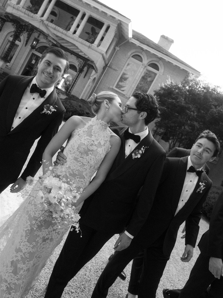Black and white photo of a bride and groom kissing, surrounded by groomsmen in tuxedos in front of a large historic house.