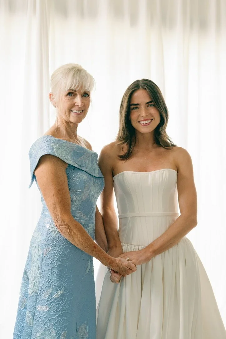 An elderly woman and a young woman, likely a bride, holding hands and smiling in front of light-colored curtains. The elderly woman wears a blue, floral lace dress, and the young woman wears a strapless white wedding gown.